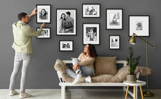Man hanging framed family photos on gray wall.