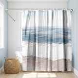 Shower curtain of soft teal sea, white surf and pale sand bands in a bright white bathroom beside a tub and basket 