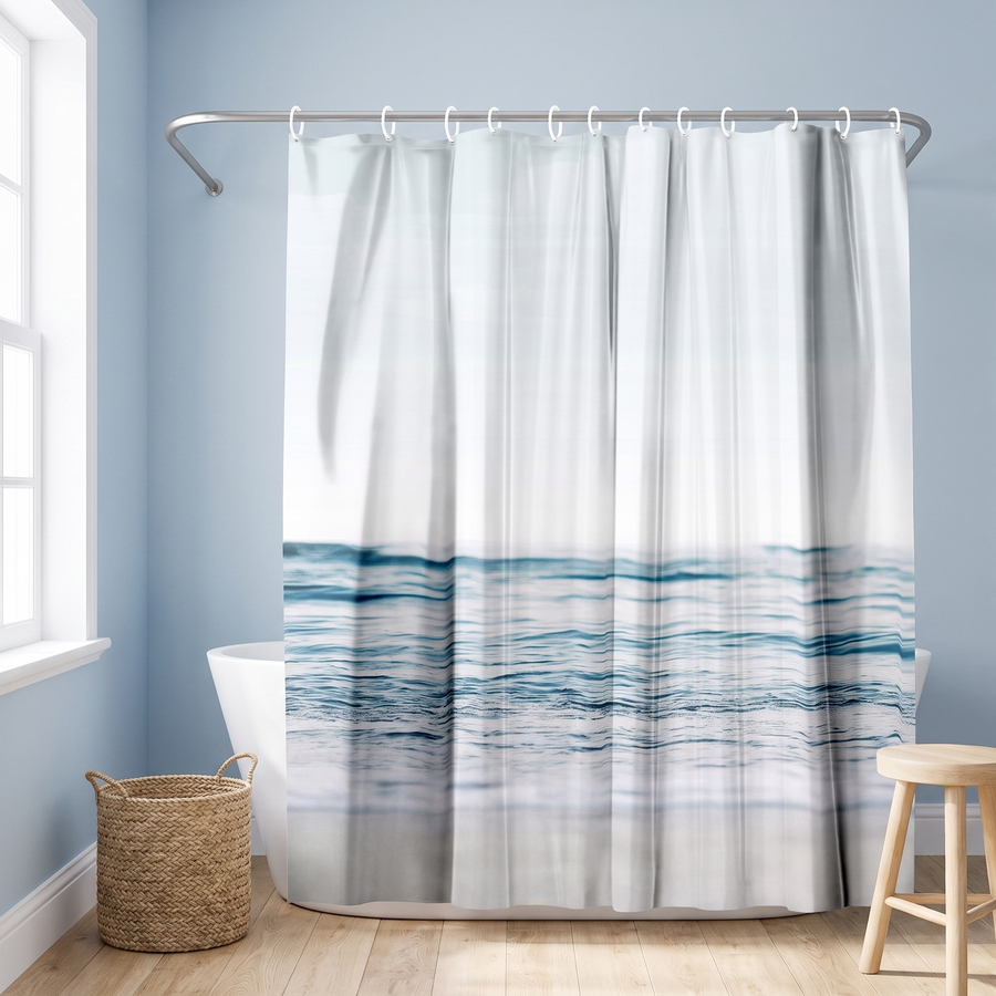 Shower curtain with blue ocean waves across pale sky and sand in a blue bathroom beside a freestanding tub and basket 