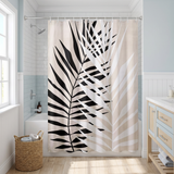 Shower curtain with a bold black palm frond on pale stripes in a bright white bathroom beside a tub and basket 