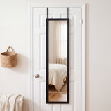 Black 12x48 over-the-door full length mirror hanging on a white bedroom door, reflecting a sunlit bed and oak floor