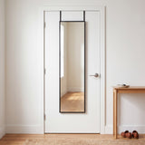 Black 12x48 over-the-door mirror on a white entryway door beside a console table, reflecting a bright hallway with oak floors