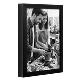 Angled black 8.5x11 deep frame, a couple potting a plant at a garden bench 