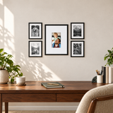 Five black frames with white mats above a walnut desk in a sunlit home office. 