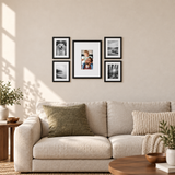 Five black frames with white mats arranged above a cream sofa, with a color digital photo in the center. 