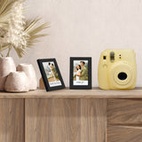 Two black Instax frames on a sideboard beside a yellow instant camera and stone vases 