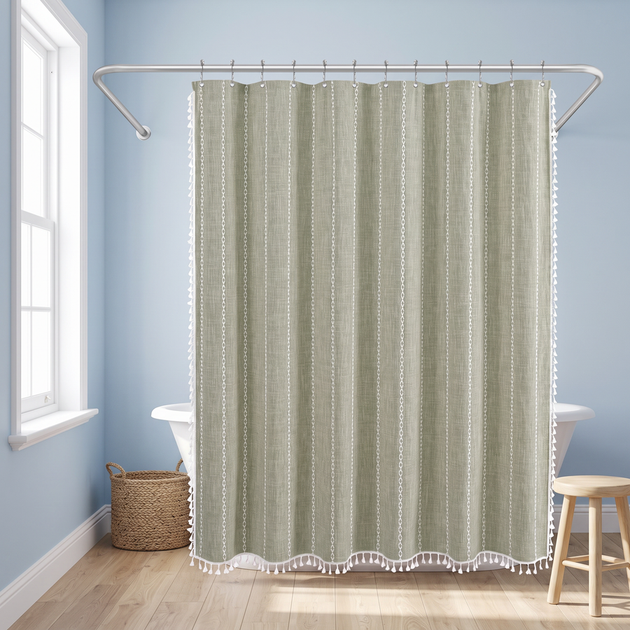 A sage green and white patterned shower curtain with white fringe trim hanging from a silver metal rod in a bright bathroom with light blue walls, a window, a basket, and a wooden stool. 