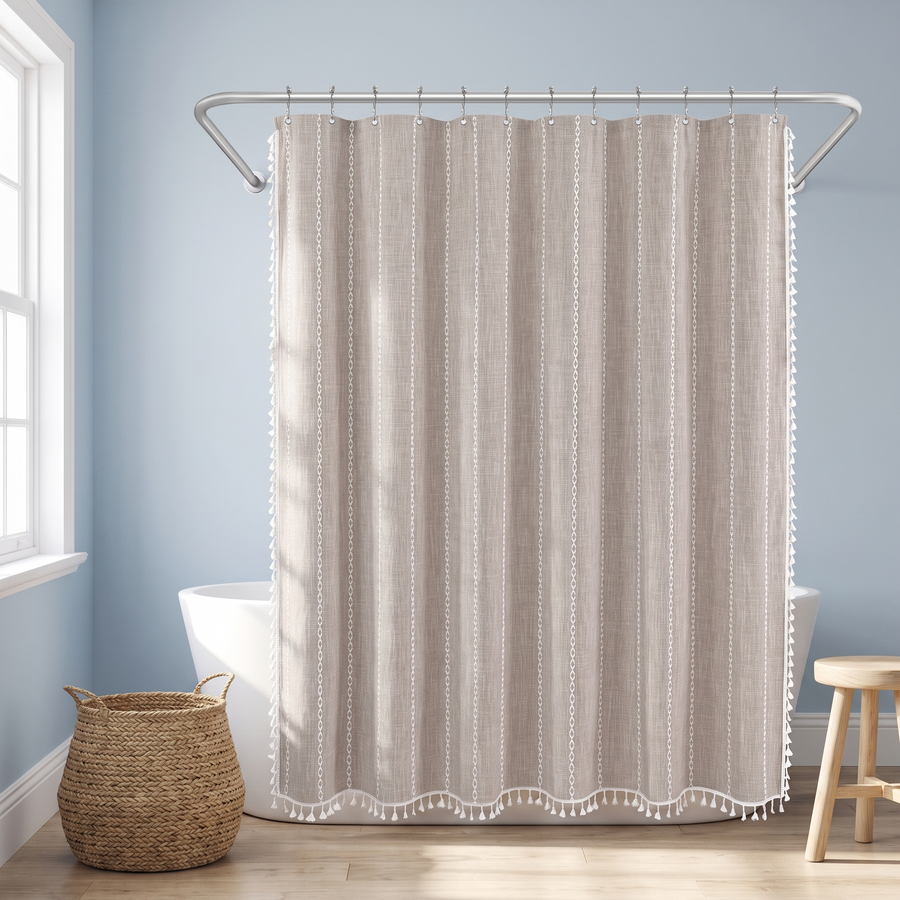 A beige shower curtain with vertical dotted stripes and white pom-pom trim hanging on a silver rod in a bright bathroom with light blue walls, a white tub, a woven basket, and a wooden stool. 