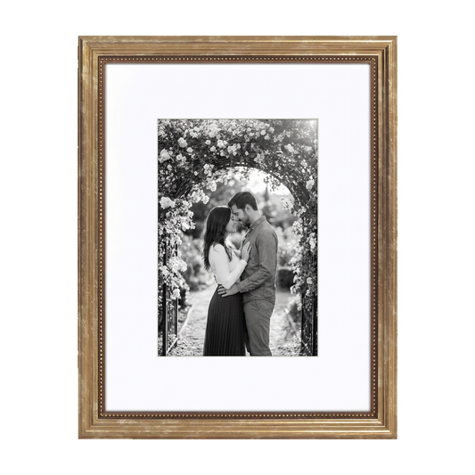 Gold beaded Heirloom frame with a wide white mat, holding a black-and-white photo of a couple beneath a floral arch
