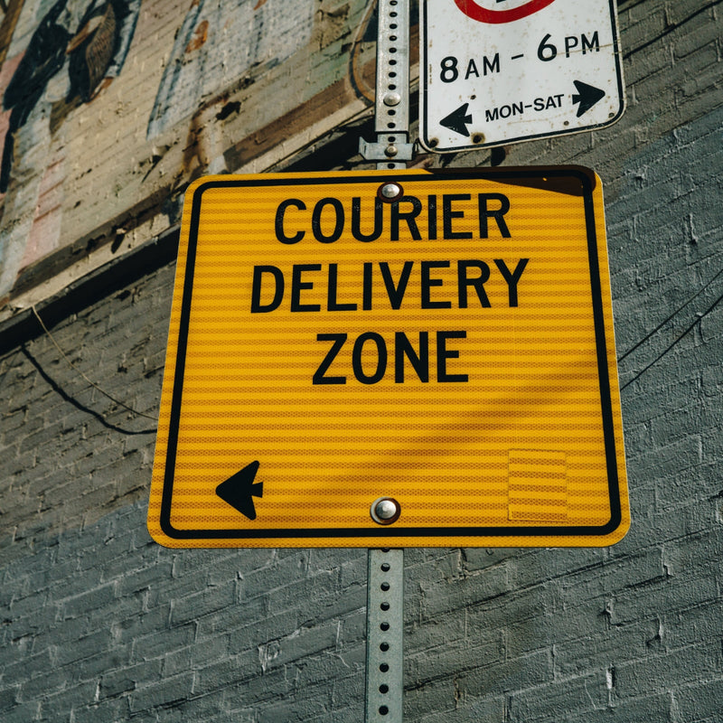 Yellow 'Courier Delivery Zone' sign on a brick wall with a smaller traffic sign above it.