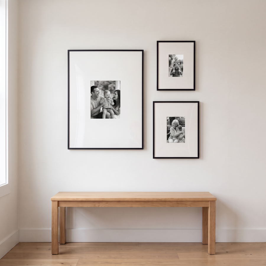 Wide Mat Mixed Trio: three black Streamline frames with wide mats: an 18x24 beside a stacked 9x12 and 11x14. Shown in an entryway, above a slim, low light-oak bench, with family photos in the frames 