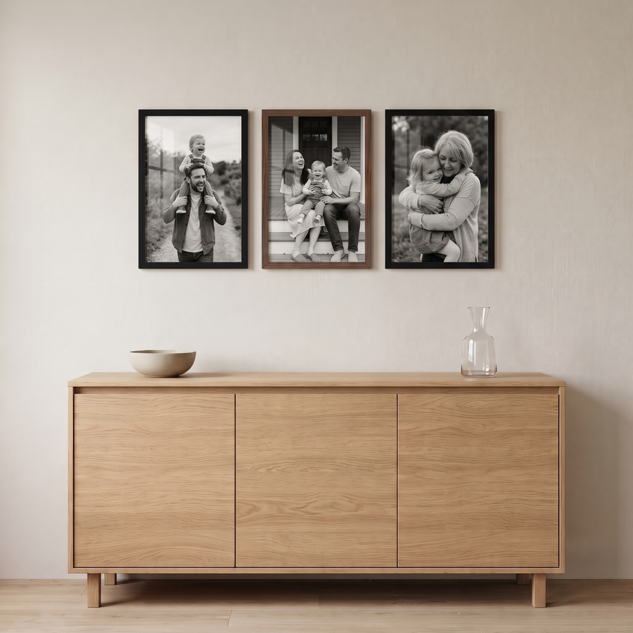 Mixed Wood Trio: three unmatted 12x18 Signature frames in a row: two black with a walnut frame in the middle. Shown in a dining room, above a light oak sideboard with a bowl and a carafe, with family photos in the frames 