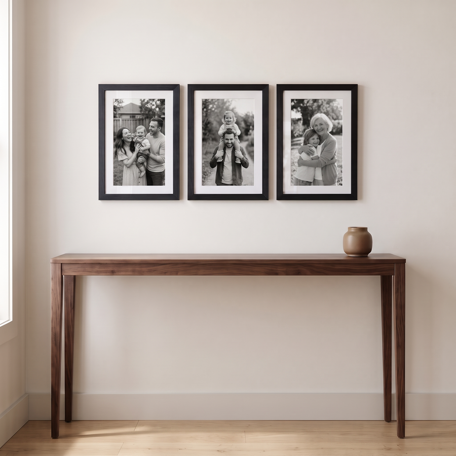 Hudson Portrait Trio: three identical black Hudson 12x18 frames, matted to 11x17, hung in one even row. Shown in an entryway, above a walnut console table with a ceramic vase, with family photos in the frames 